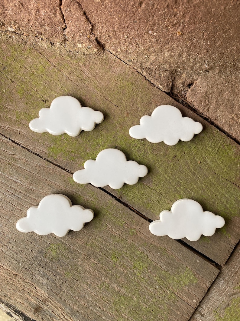 May include: Five white ceramic cloud-shaped ornaments on a wooden surface. The clouds are all the same size and shape, and they are arranged in a random pattern.