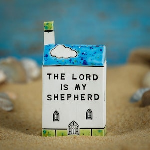 Puede incluir: Una caja de cerámica blanca con forma de iglesia con techo azul y chimenea. El texto "THE LORD IS MY SHEPHERD" se muestra en la parte delantera. La caja tiene un diseño de nube y pequeños detalles de ventana. La base tiene un borde verde.