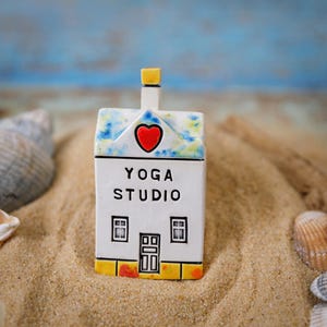 Può includere: Una piccola casa in ceramica bianca con un cuore rosso e le parole "YOGA STUDIO" sulla facciata. La casa è seduta su un letto di sabbia.