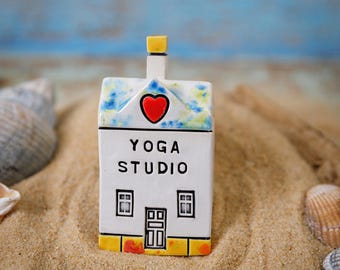 以下が含まれることがあります： 赤いハートと「YOGA STUDIO」の文字が書かれた小さな白い陶器の家。家は砂の上に置かれています。