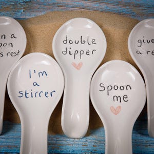 May include: White ceramic spoons with handwritten phrases like "double dipper" and "spoon me" are arranged on a sandy surface. Each spoon has a unique message, some with small heart drawings. The spoons rest on a blue wooden surface.