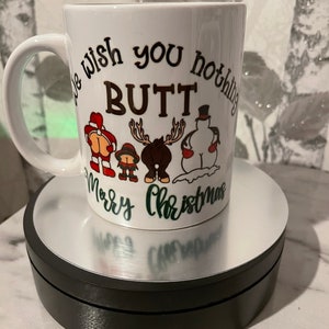 May include: White ceramic mug with a black and green design. The mug has a handle and says "We wish you nothing BUTT Merry Christmas" with a cartoon Santa, snowman, reindeer, and elf.