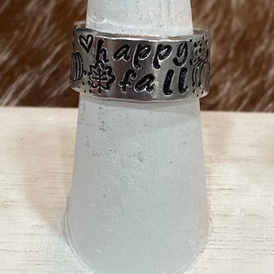 May include: Silver-toned ring with the words "happy fall" etched into the band, along with leaf and pumpkin designs. The ring is displayed on a white ring holder, showcasing its fall-themed design.