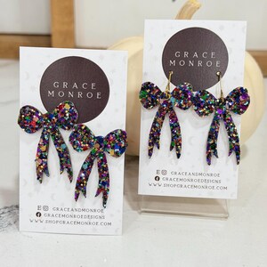 May include: Glittery bow earrings in shades of purple, green, and gold. The earrings are displayed on white cards with the text "GRACE MONROE" and social media information. The bows have a textured, sparkly appearance.