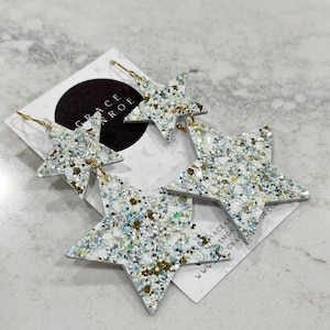 May include: A pair of silver glitter star earrings with gold accents. The earrings are layered with two stars on each side.