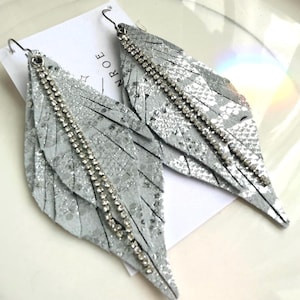 May include: A pair of silver-colored feather-shaped earrings. Each earring features a textured, glittery surface and is adorned with a line of small, sparkling rhinestones. The earrings hang from silver-colored hooks.