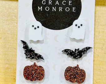 May include: Halloween-themed stud earrings featuring a white ghost, a black bat with glitter, and a glittery orange pumpkin. The card includes the text "GRACE MONROE" and social media information.