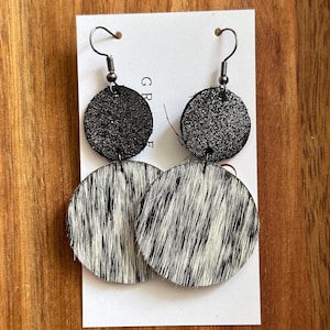 May include: A pair of dangle earrings with two round discs. The top disc is black with a subtle sparkle and the bottom disc is white with black cowhide print.