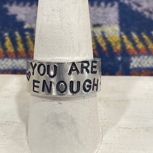 May include: A silver ring with the words "You Are Enough" engraved on it. The ring is on a white display stand.