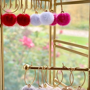 May include: Gold hoop earrings with fluffy pom poms in various colors, including pink, white, and brown. The earrings are displayed on a gold earring holder.