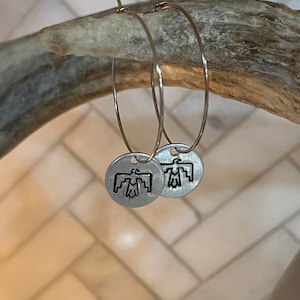May include: Silver hoop earrings with a small, round charm featuring a stylized bird design.