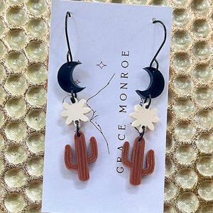 May include: A pair of dangle earrings featuring a black crescent moon, a white flower, and a brown cactus charm. The earrings are on a white card with the text "GRACE MONROE" printed on it.