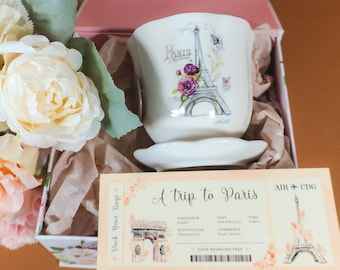 May include: A white ceramic teacup and saucer with a floral design and an image of the Eiffel Tower. The teacup and saucer are in a pink gift box with a white tissue paper lining. A boarding pass with the text "A trip to Paris" is on top of the box.
