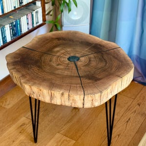 Op de afbeelding: Houten bijzettafel met een natuurlijke rand. Het lichtbruine tafelblad toont de natuurlijke houtnerf en donkere accenten. De tafel staat op drie zwarte hairpin poten, wat een moderne, rustieke uitstraling geeft. De diameter van de tafel is ongeveer 61 cm.