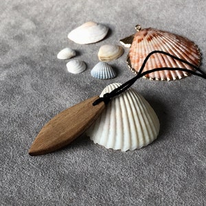 Pode incluir: Um pingente de madeira em forma de folha, pendurado em um cordão preto com um charme de concha. O pingente é cercado por várias conchas pequenas em um fundo cinza.