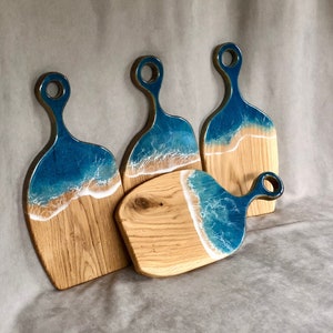 May include: Set of four wooden cutting boards with blue and white resin ocean wave designs. The boards have a rounded shape and a handle.