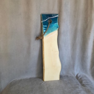 May include: A long, wooden chopping board with a blue and white epoxy resin inlay. The board has a hole for hanging and a rope attached to it.