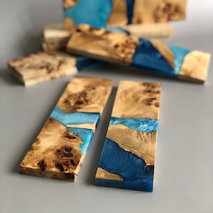 Può includere: Diversi blocchi di legno rettangolari con una venatura marrone chiaro e strutturata sono intervallati da resina blu vibrante. La resina riempie gli spazi vuoti e crea un contrasto sorprendente. I blocchi sono disposti su una superficie grigia neutra.