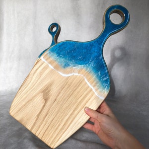 May include: A wooden cutting board with a blue and white resin ocean wave design. The board has a handle with a hole for hanging.