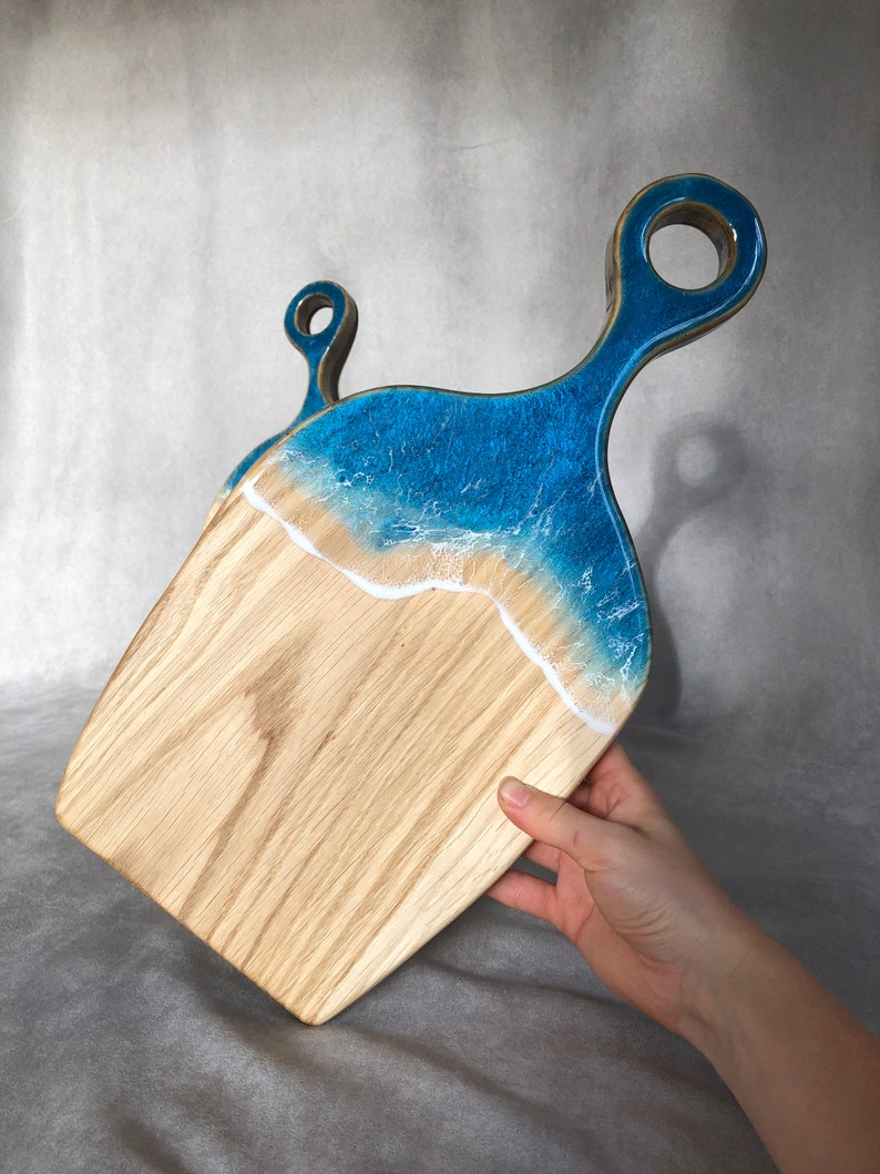 May include: A wooden cutting board with a blue and white resin ocean wave design. The board has a handle with a hole for hanging.