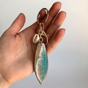 May include: A gold key chain with a wooden surfboard charm featuring a blue and white wave design. The charm is accented with a small white seashell.