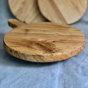 May include: A round wooden chopping board with a handle. The board is made of light-coloured wood and has a natural grain pattern. Two additional boards with blue and white resin accents are visible in the background.