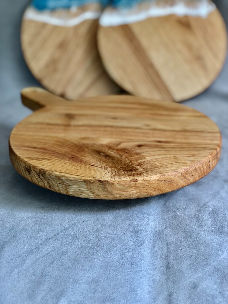 May include: A round wooden chopping board with a handle. The board is made of light-coloured wood and has a natural grain pattern. Two additional boards with blue and white resin accents are visible in the background.