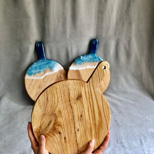 May include: Three wooden serving boards. The front board is round with a handle and a natural wood grain. Two boards in the background have a blue and white resin design resembling a beach scene. The handles are a dark blue colour.