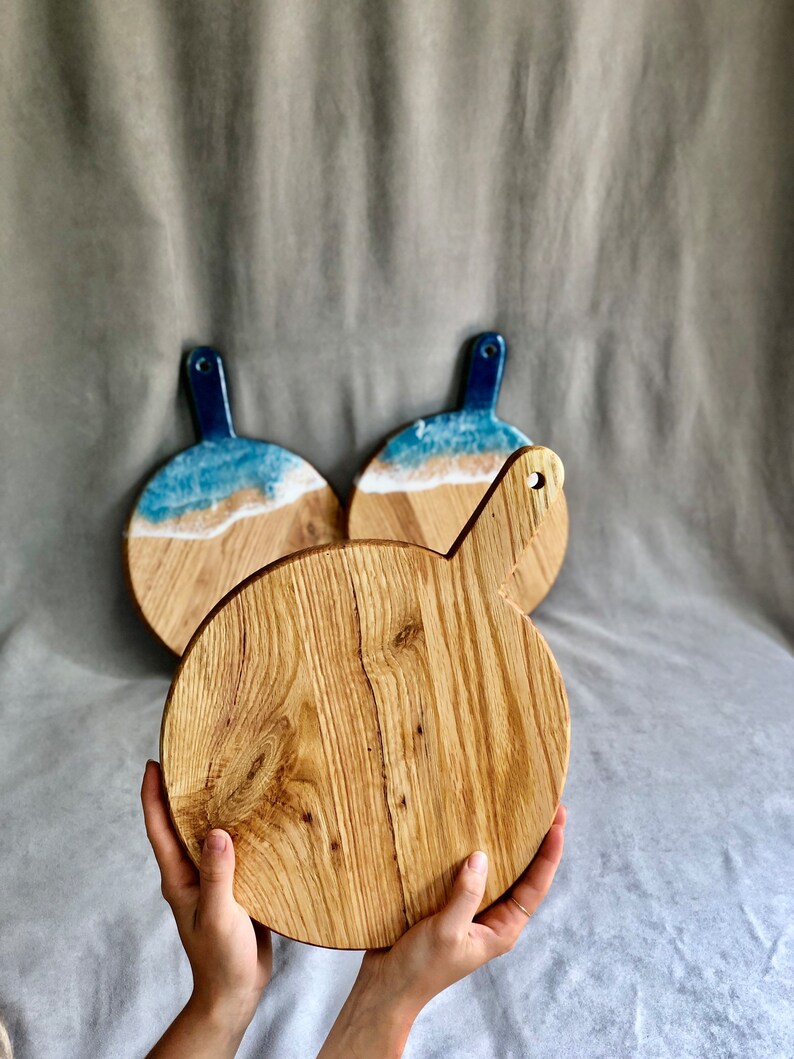 May include: Three wooden serving boards. The front board is round with a handle and a natural wood grain. Two boards in the background have a blue and white resin design resembling a beach scene. The handles are a dark blue colour.