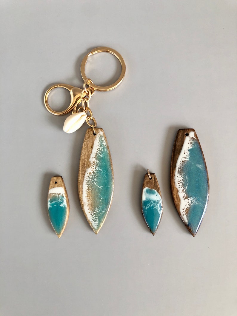 May include: Set of three wooden surfboard keychains with blue and white resin ocean wave designs. The largest keychain has a gold chain and a small seashell charm.