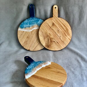 May include: Three round wooden chopping boards with handles. Two boards feature a blue and white resin design resembling ocean waves. The third board has a similar design. The handles are dark blue.