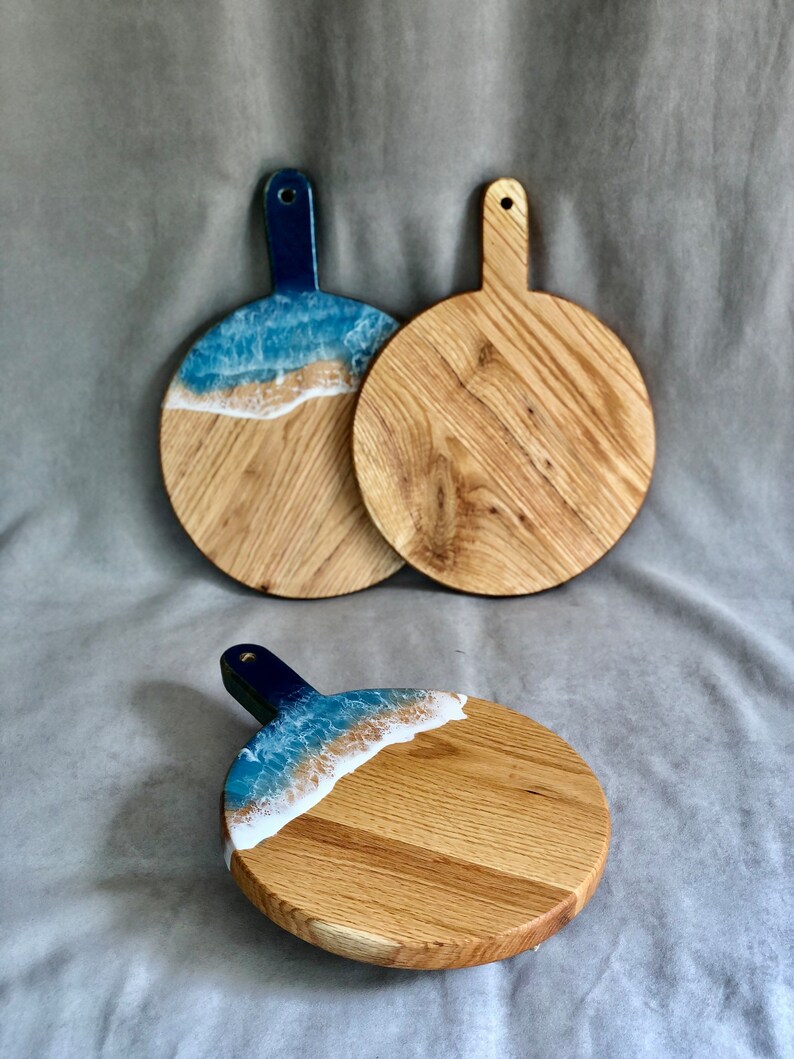 May include: Three round wooden chopping boards with handles. Two boards feature a blue and white resin design resembling ocean waves. The third board has a similar design. The handles are dark blue.