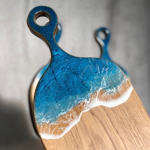 May include: A wooden cutting board with a blue and white epoxy resin design that resembles an ocean wave. The board has a handle with a hole in it.