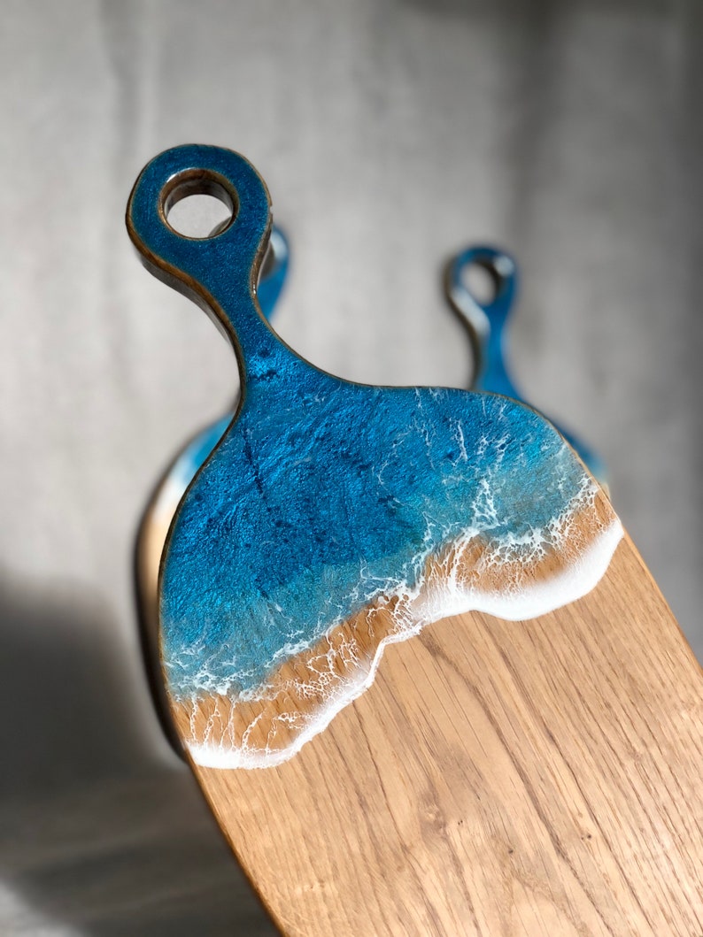 May include: A wooden cutting board with a blue and white epoxy resin design that resembles an ocean wave. The board has a handle with a hole in it.