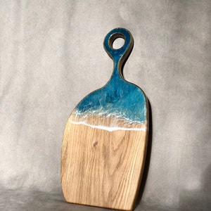 May include: A wooden cutting board with a blue and white resin inlay that resembles ocean waves. The board has a handle with a hole in it.
