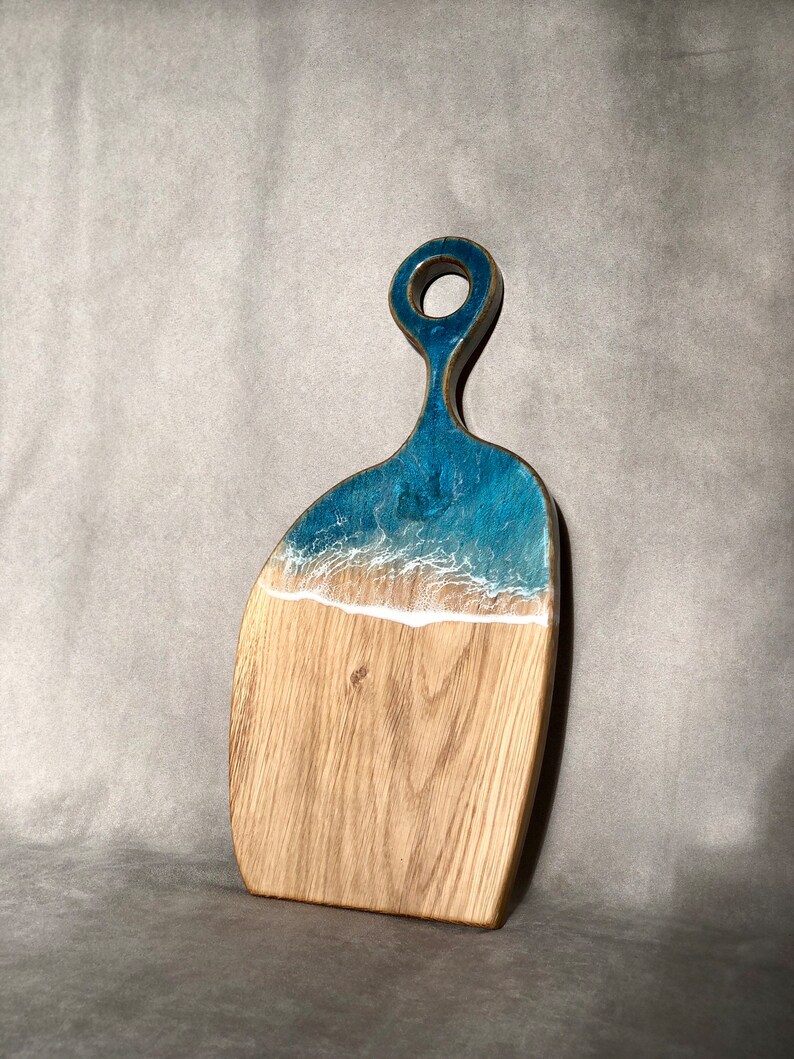 May include: A wooden cutting board with a blue and white resin inlay that resembles ocean waves. The board has a handle with a hole in it.