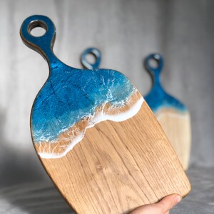 May include: A wooden cutting board with a blue and white resin ocean wave design. The board has a handle and is perfect for serving cheese, charcuterie, or other appetizers.