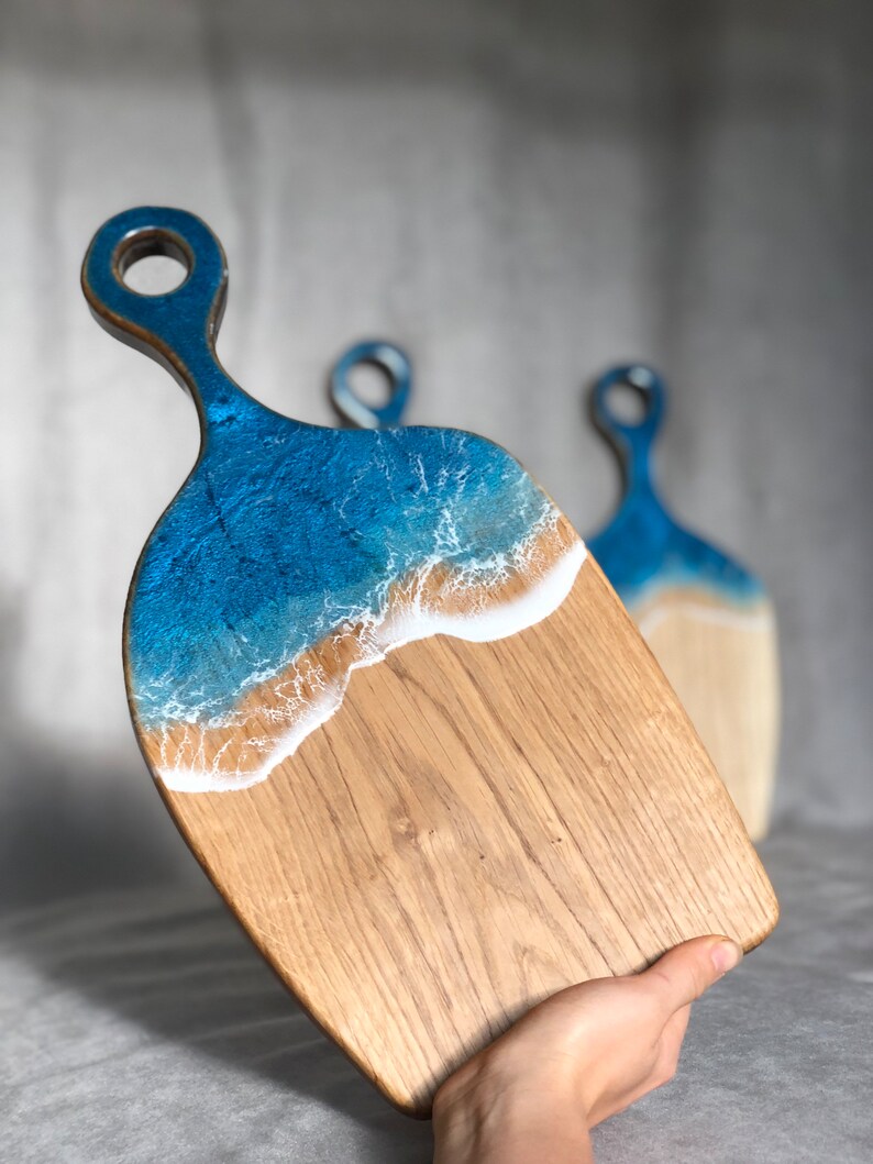 May include: A wooden cutting board with a blue and white resin ocean wave design. The board has a handle and is perfect for serving cheese, charcuterie, or other appetizers.