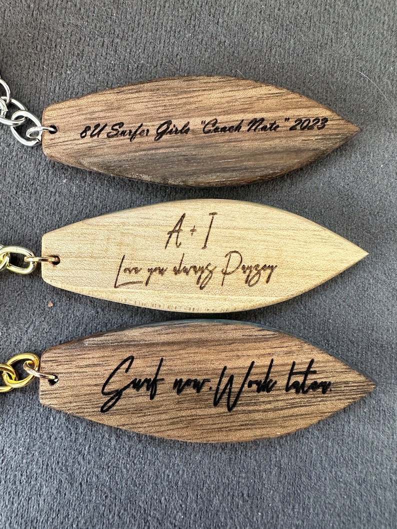 May include: Three wooden surfboard-shaped keychains with different engraved messages. The top keychain is dark brown and reads "8U Surfer Girls "Coach Nate" 2023". The middle keychain is light brown and reads "A+I Love you always Paige". The bottom keychain is dark brown and reads "Surf now. Work later."