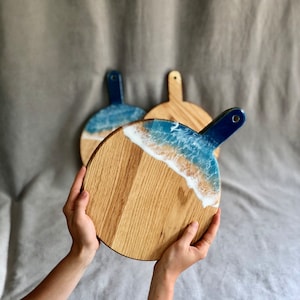 May include: Round wooden serving boards with a handle. The boards have a blue and white resin design that looks like ocean waves. The wood has a natural grain, and the handles are painted blue. The boards are held by a person.