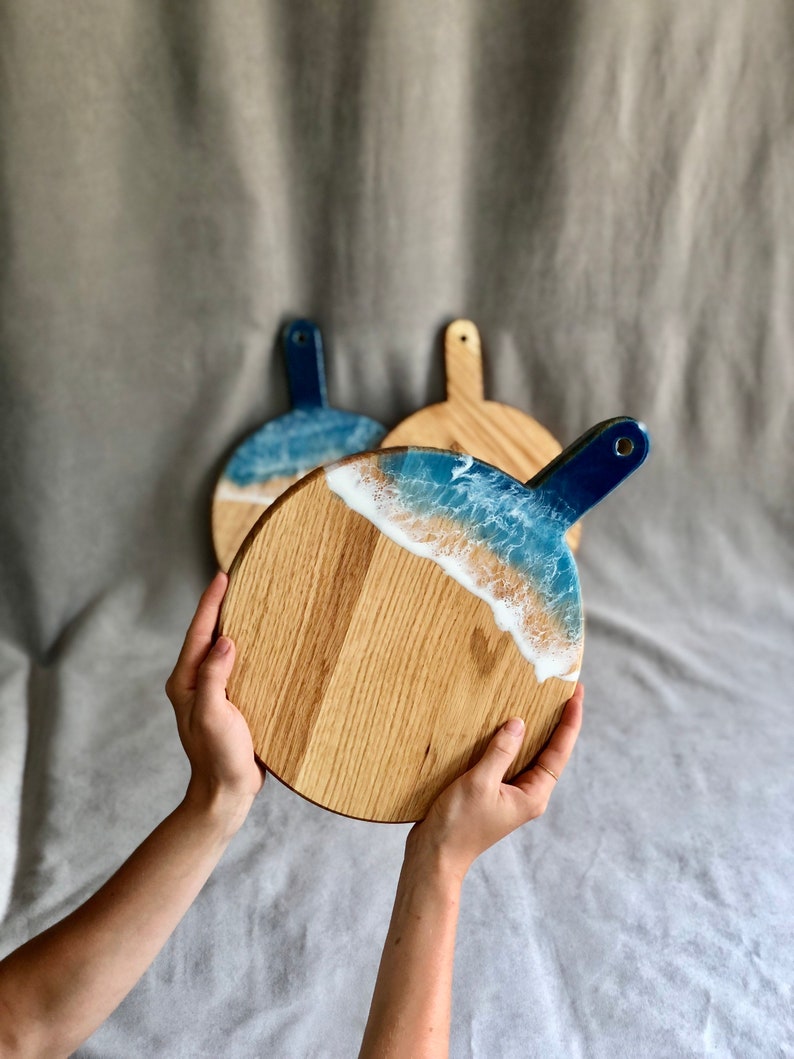 May include: Round wooden serving boards with a handle. The boards have a blue and white resin design that looks like ocean waves. The wood has a natural grain, and the handles are painted blue. The boards are held by a person.