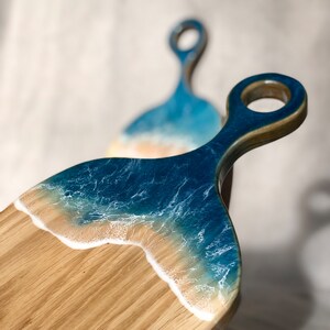 May include: A wooden cutting board with a blue and white resin ocean wave design. The board has a handle with a loop at the end.