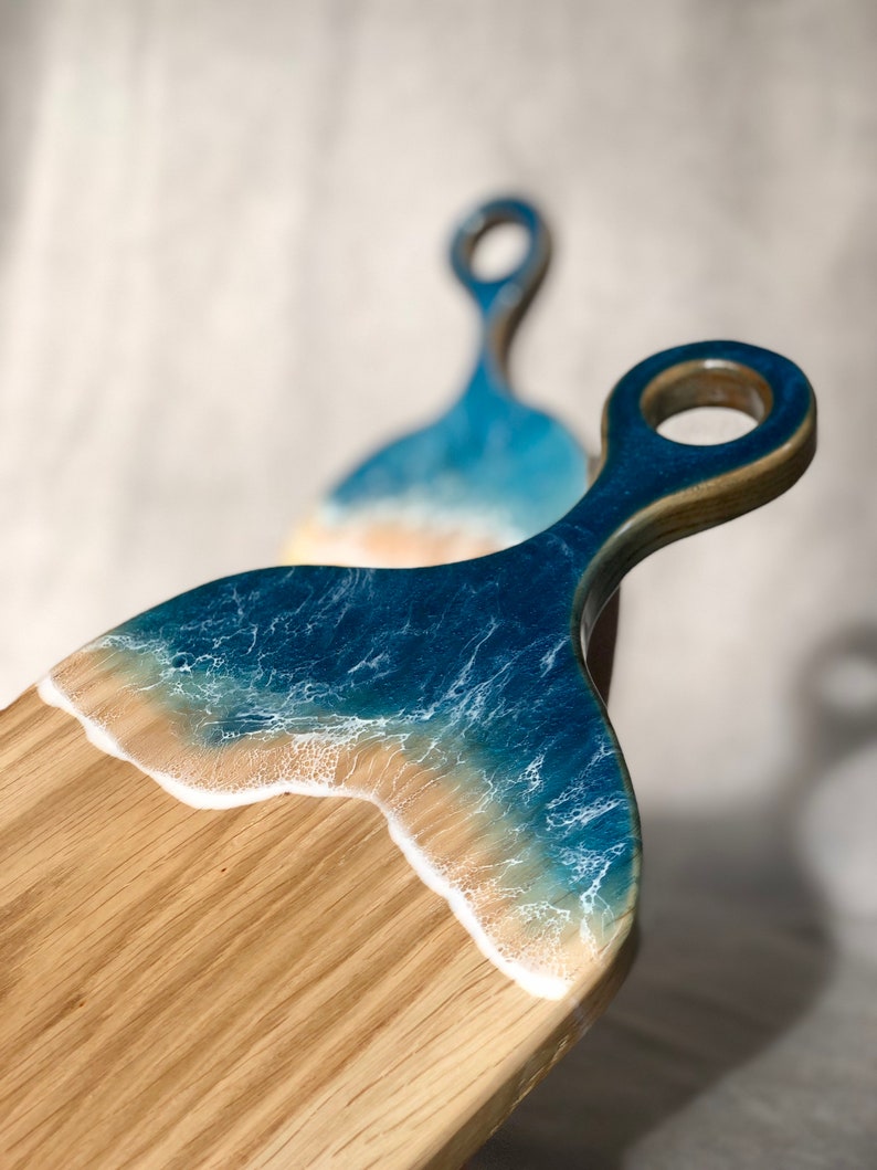 May include: A wooden cutting board with a blue and white resin ocean wave design. The board has a handle with a loop at the end.