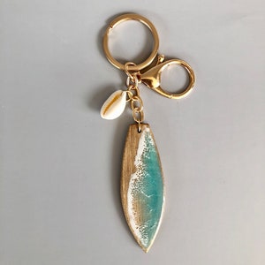 May include: Gold key chain with a surfboard charm featuring a blue and white wave design and a small white seashell charm.