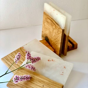Puede incluir: Conjunto de cuatro posavasos de resina blanca con motas rojas, cada uno con una base de madera. Los posavasos est&aacute;n apilados en un soporte de madera.