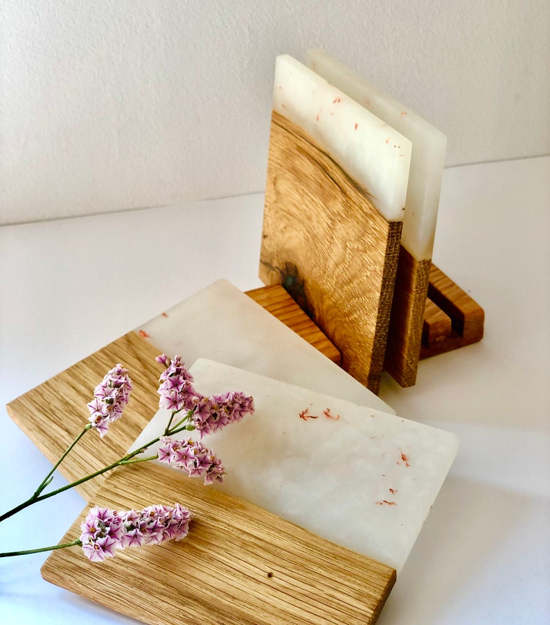 Puede incluir: Conjunto de cuatro posavasos de resina blanca con motas rojas, cada uno con una base de madera. Los posavasos est&aacute;n apilados en un soporte de madera.