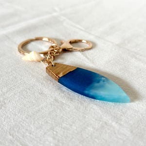 Könnte beinhalten: Ein blauer und weißer Schlüsselanhänger aus Harz in Form eines Surfbretts mit einem goldfarbenen Metallring und einem kleinen Muschel-Anhänger.