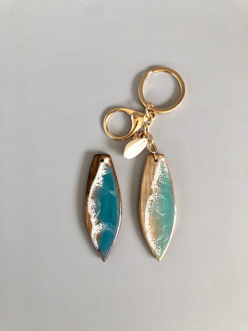 May include: Two wooden surfboard keychains with blue and white resin waves. The keychains are attached to a gold chain with a gold lobster clasp and a small white seashell charm.