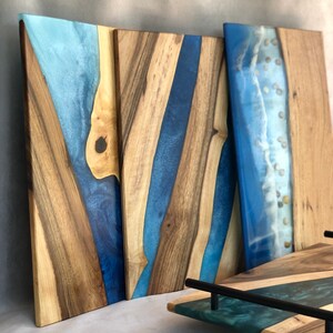 May include: Three wooden serving boards with blue and teal epoxy resin. The boards have a natural wood grain and are rectangular in shape. The epoxy resin creates a river-like design on the boards. The boards are perfect for serving cheese, charcuterie, or other appetizers.
