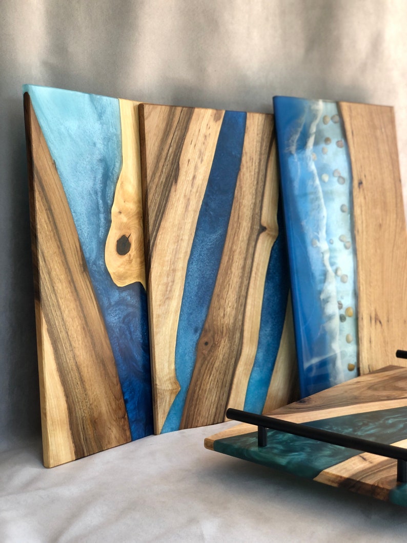 May include: Three wooden serving boards with blue and teal epoxy resin. The boards have a natural wood grain and are rectangular in shape. The epoxy resin creates a river-like design on the boards. The boards are perfect for serving cheese, charcuterie, or other appetizers.
